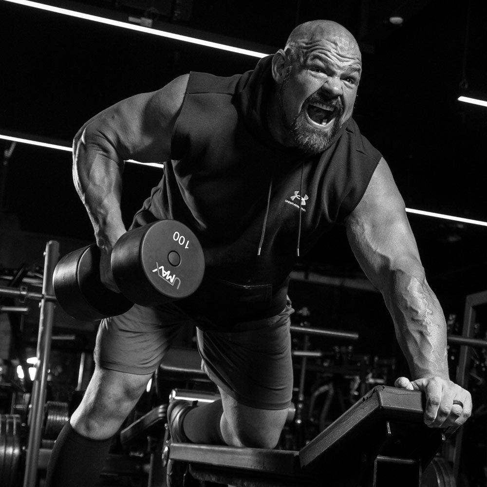 Brian Shaw in a black sleeveless hoodie and shorts lifts a 100 kg dumbbell in a gym setting, captured in dramatic black and white.