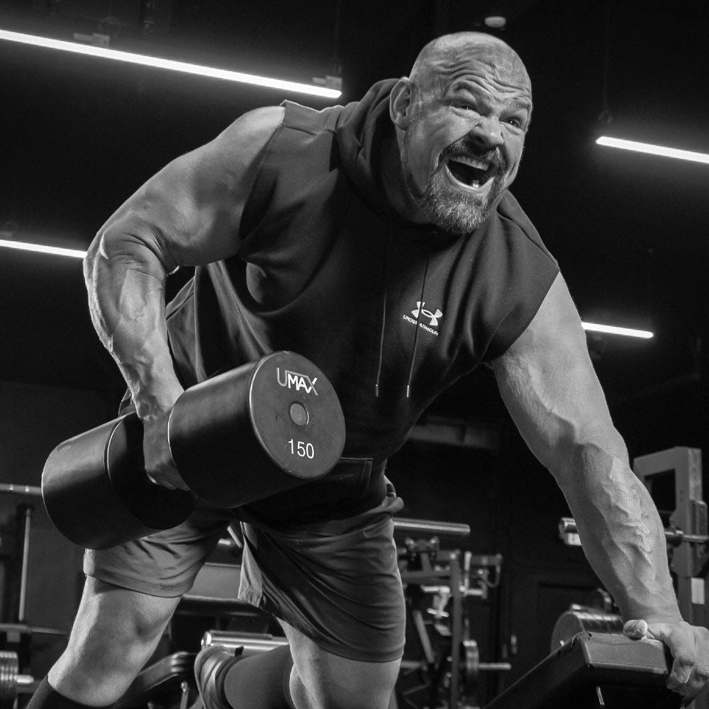 Brian Shaw in a black sleeveless hoodie and shorts lifts a 150 lb dumbbell in a gym setting, captured in dramatic black and white.