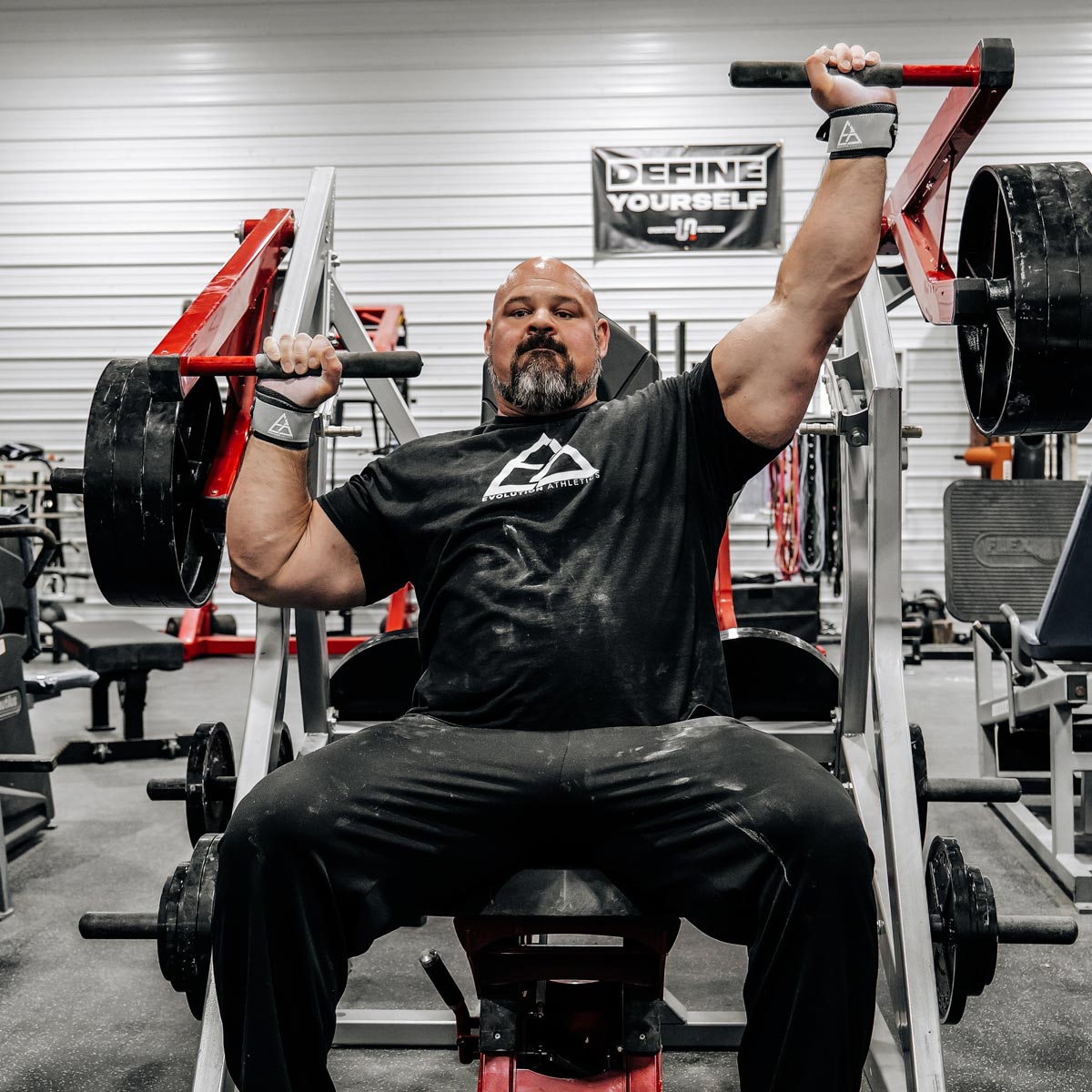 Brian Shaw performing a bench press in a gym setting with weights and gym equipment around.