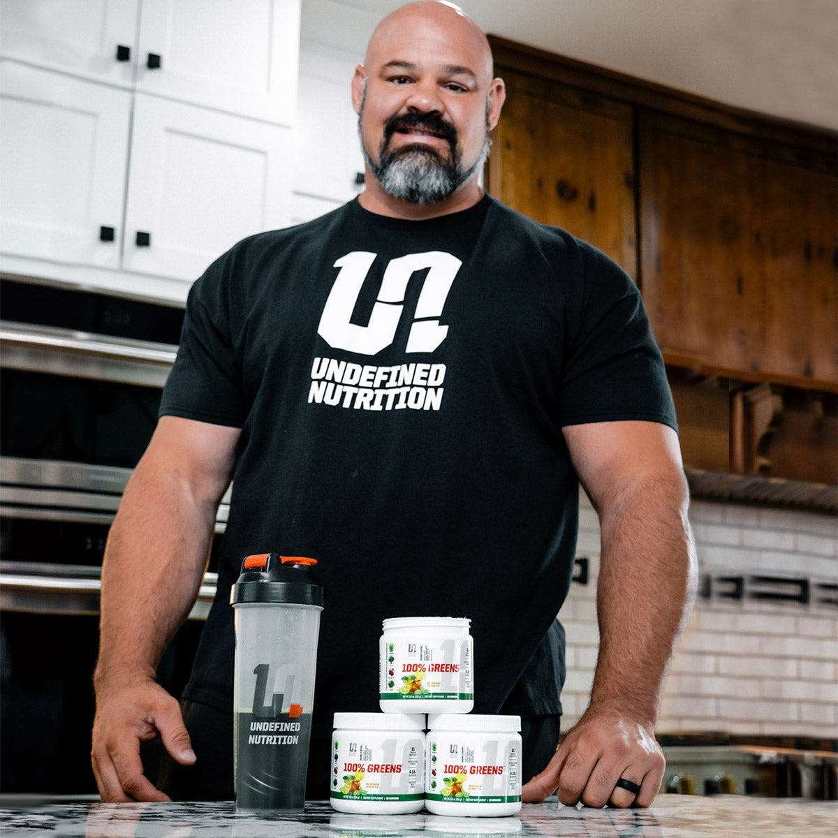 Brian Shaw wearing a black 'Undefined Nutrition' t-shirt with nutritional supplements on a kitchen counter.