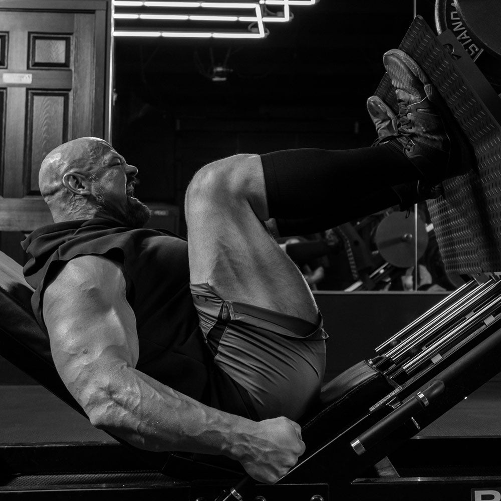 Brian Shaw in a gym performing a leg extension exercise, shown in black and white. He is wearing a sleeveless shirt, shorts, and knee sleeves, pushing a weight stack with his legs extended.