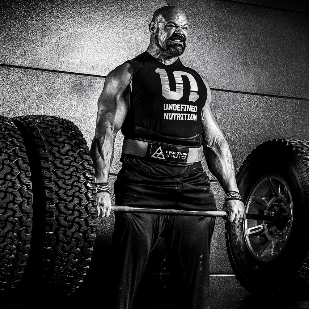Brian Shaw in a black sleeveless tank top with 'UNDEFINED NUTRITION' printed on the front, lifting a barbell on a gym floor between large tires. He is wearing a belt with the Evolution Athletics lifting belt.