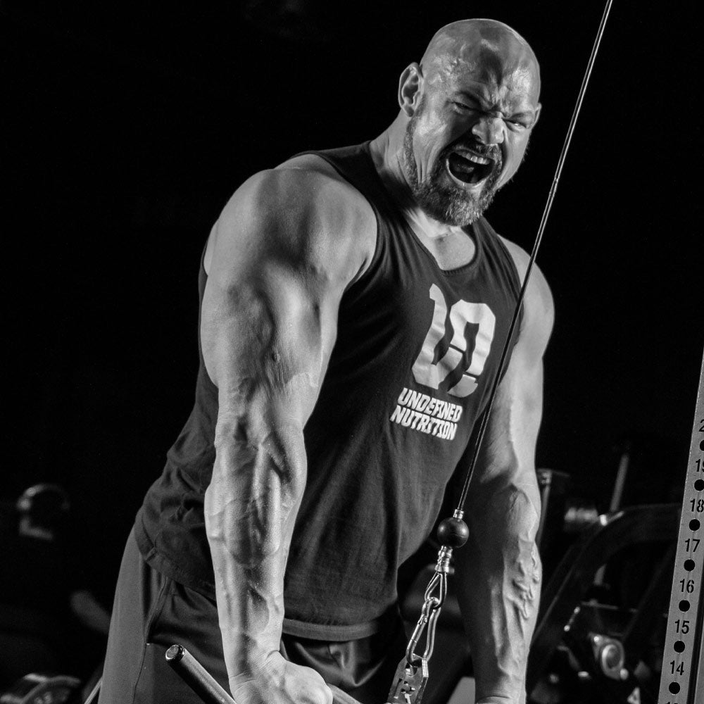 Brian Shaw in a gym wearing a black Undefined Nutrition tank top, performing a cable tricep pushdown against a dark background.
