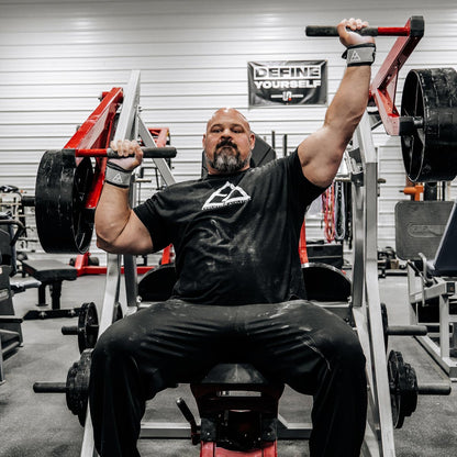 Brian Shaw performing a bench press in a gym setting with weights and gym equipment around.