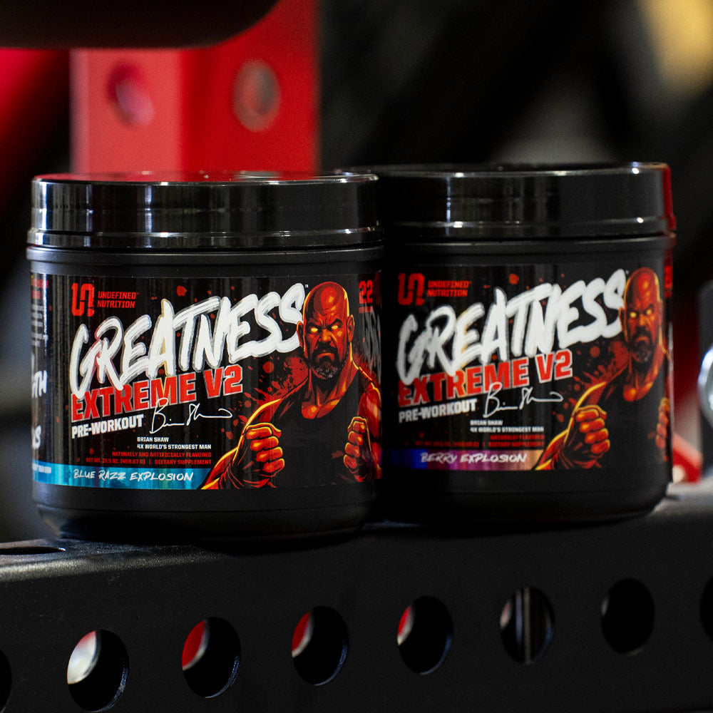 Two black tubs of Undefined Nutrition Greatness V2 Extreme Pre-Workout powder on a black metal shelf in a gym setting.