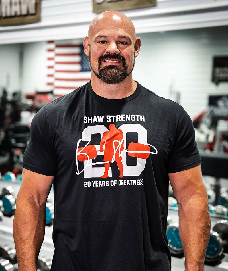 Brian Shaw stands in a gym wearing a black t-shirt with a white and orange graphic that reads 'SHAW STRENGTH 20 YEARS OF GREATNESS'.